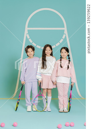 Three kids stand together holding tennis rackets in a bright blue space. They smile and wear sports clothes. Tennis balls are scattered on the ground around them. Three kids stand together holding tennis rackets in a bright blue space. They smile and wear sports clothes. Tennis balls are scattered on the ground around them. 139259622