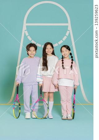 Three children stand together, holding tennis rackets. They wear colorful clothing and smile at the camera. The background features a tennis net design. Three children stand together, holding tennis rackets. They wear colorful clothing and smile at the camera. The background features a tennis net design. 139259623