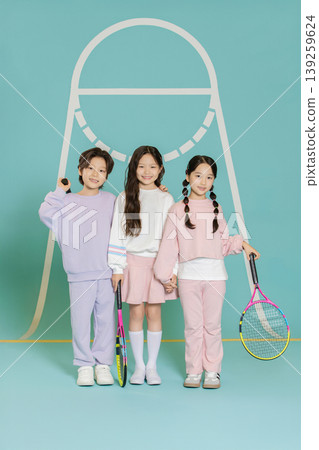 Three children stand together in a studio, each holding a tennis racket. They wear casual outfits in different colors and have a friendly pose. 139259624