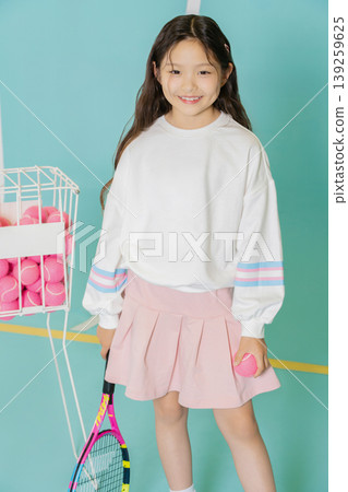 A young girl smiles while holding a tennis ball in her hand. She stands near a basket filled with pink tennis balls. The indoor court has blue walls and a simple design. A young girl smiles while holding a tennis ball in her hand. She stands near a basket filled with pink tennis balls. The indoor court has blue walls and a simple design. 139259625