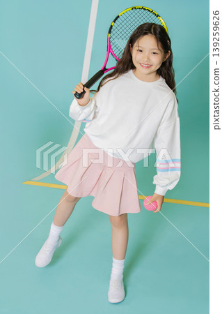 A girl holds a tennis racket in one hand and a tennis ball in the other while standing on a light blue indoor court. She is smiling and wearing casual sports clothes. A girl holds a tennis racket in one hand and a tennis ball in the other while standing on a light blue indoor court. She is smiling and wearing casual sports clothes. 139259626
