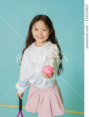 A girl stands smiling while holding a tennis ball in one hand and a racket in the other. She wears a light sweater and a skirt, ready for a fun time playing tennis. A girl stands smiling while holding a tennis ball in one hand and a racket in the other. She wears a light sweater and a skirt, ready for a fun time playing tennis. 139259627