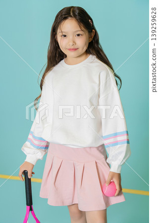 A young girl stands holding a tennis racket and a pink ball. She wears a white shirt and a pink skirt. The background is blue and she looks cheerful. This scene takes place indoors during the day. A young girl stands holding a tennis racket and a pink ball. She wears a white shirt and a pink skirt. The background is blue and she looks cheerful. This scene takes place indoors during the day. 139259628