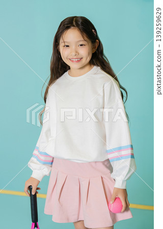A young girl stands smiling while holding a tennis racket in one hand and a pink ball in the other. She wears a white shirt and a pink skirt. A young girl stands smiling while holding a tennis racket in one hand and a pink ball in the other. She wears a white shirt and a pink skirt. 139259629