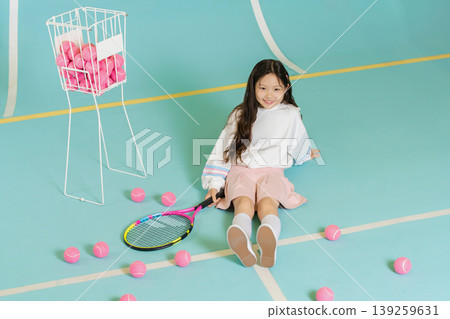 A girl sits on a tennis court surrounded by pink tennis balls. She holds a racket and looks happy. A white basket holds more balls nearby. It is a sunny afternoon, perfect for practice. A girl sits on a tennis court surrounded by pink tennis balls. She holds a racket and looks happy. A white basket holds more balls nearby. It is a sunny afternoon, perfect for practice. 139259631