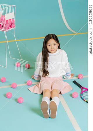 A girl is sitting on the tennis court surrounded by pink tennis balls. She has a racket beside her and is wearing a white sweater and pink skirt. The setting is bright and simple. A girl is sitting on the tennis court surrounded by pink tennis balls. She has a racket beside her and is wearing a white sweater and pink skirt. The setting is bright and simple. 139259632