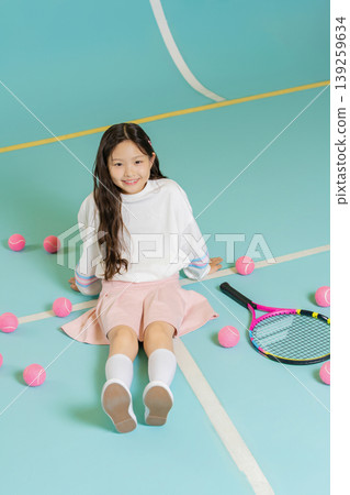 A girl sits on a tennis court with pink tennis balls scattered around her. She wears a sweater and skirt while having a fun moment with her racket nearby. A girl sits on a tennis court with pink tennis balls scattered around her. She wears a sweater and skirt while having a fun moment with her racket nearby. 139259634