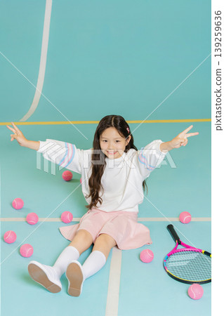 A child sits on a blue tennis court surrounded by pink tennis balls. She smiles and makes a peace sign with her fingers. A tennis racket lies beside her as she enjoys the activity. A child sits on a blue tennis court surrounded by pink tennis balls. She smiles and makes a peace sign with her fingers. A tennis racket lies beside her as she enjoys the activity. 139259636