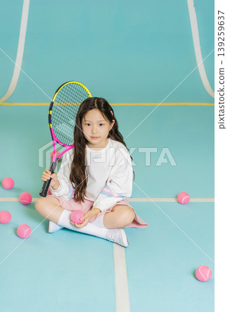 A young girl sits on the tennis court holding a racket with pink tennis balls around her. She focuses on her training while preparing for her next move in the sport. A young girl sits on the tennis court holding a racket with pink tennis balls around her. She focuses on her training while preparing for her next move in the sport. 139259637