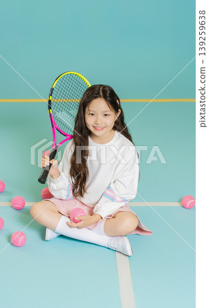 A girl sits on the floor of a tennis court holding a racket. Around her are pink tennis balls. She is smiling and wearing a white top with a skirt. This scene shows practice time. A girl sits on the floor of a tennis court holding a racket. Around her are pink tennis balls. She is smiling and wearing a white top with a skirt. This scene shows practice time. 139259638