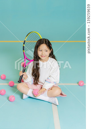 A young girl is sitting on a blue gym court with pink tennis balls around her. She holds a tennis racket and smiles, enjoying her time playing the sport. A young girl is sitting on a blue gym court with pink tennis balls around her. She holds a tennis racket and smiles, enjoying her time playing the sport. 139259639