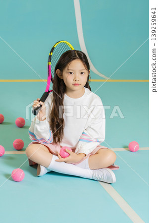 A young girl sits on a blue sports court with tennis balls around her. She holds a racket in one hand and a ball in the other. She looks serious and ready to play tennis. A young girl sits on a blue sports court with tennis balls around her. She holds a racket in one hand and a ball in the other. She looks serious and ready to play tennis. 139259641