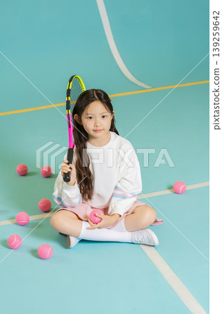 A young child sits cross-legged on a tennis court. She holds a racket in one hand and a tennis ball in the other. Pink tennis balls are scattered around her. The setting is bright and inviting. A young child sits cross-legged on a tennis court. She holds a racket in one hand and a tennis ball in the other. Pink tennis balls are scattered around her. The setting is bright and inviting. 139259642