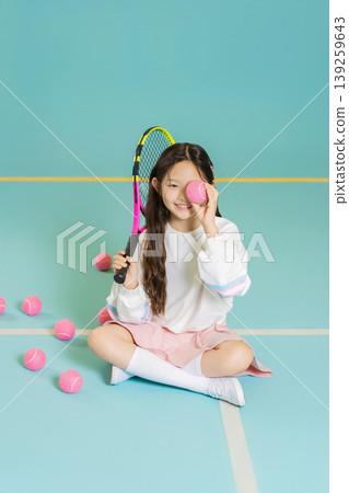 A girl sits on a tennis court, holding a pink tennis ball to her eye. She smiles as she plays among scattered pink tennis balls. It is a bright and fun setting for tennis. A girl sits on a tennis court, holding a pink tennis ball to her eye. She smiles as she plays among scattered pink tennis balls. It is a bright and fun setting for tennis. 139259643