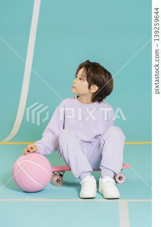 A child sits on a skateboard holding a pink basketball in a bright room. The child wears purple clothes and white shoes. The background has a blue color and white lines on the ground. A child sits on a skateboard holding a pink basketball in a bright room. The child wears purple clothes and white shoes. The background has a blue color and white lines on the ground. 139259644
