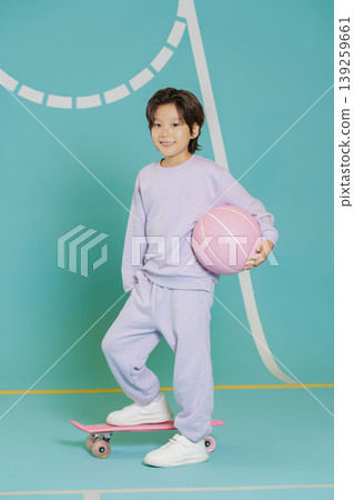 A child stands on a skateboard while holding a pink basketball in a brightly colored indoor space. The background shows a sports court with lines. It is a fun time for play. A child stands on a skateboard while holding a pink basketball in a brightly colored indoor space. The background shows a sports court with lines. It is a fun time for play. 139259661