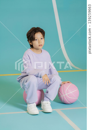 A child sits on a basketball court with two pink basketballs. The background features bright colors and clean lines. The child wears a casual outfit and seems focused. A child sits on a basketball court with two pink basketballs. The background features bright colors and clean lines. The child wears a casual outfit and seems focused. 139259663
