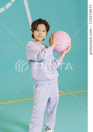 A child stands in a gym holding a pink basketball. The court has a blue background and yellow lines. The child is wearing a purple outfit. They appear to be ready for practice. A child stands in a gym holding a pink basketball. The court has a blue background and yellow lines. The child is wearing a purple outfit. They appear to be ready for practice. 139259675