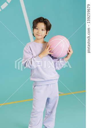 A child stands in a gym holding a pink basketball. They wear a matching outfit and smile during practice time. The court has a blue background with markings. 139259679