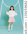 A girl stands on a gym floor holding a skipping rope. She wears a white sweatshirt and a pink skirt. The area has blue walls with markings and shows a fun activity for children. 139259682