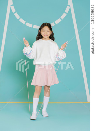 A girl stands on a gym floor holding a skipping rope. She wears a white sweatshirt and a pink skirt. The area has blue walls with markings and shows a fun activity for children. 139259682
