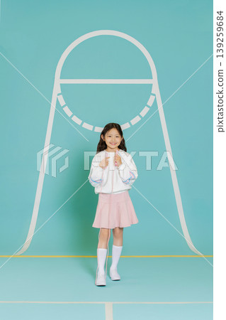 A young girl is smiling while standing against a blue background with simple shapes. She wears a white top and a pink skirt, looking happy. The scene is bright and cheerful, showcasing a fun moment. 139259684