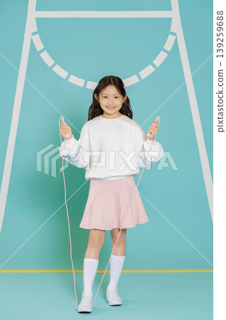 A girl stands in a bright setting and holds a skipping rope in each hand. She wears a white top, a pink skirt, and white socks. She appears happy and ready to play. 139259688