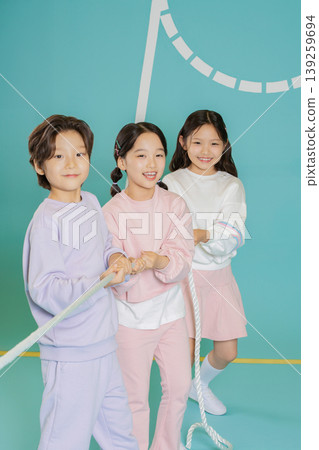 Three kids are engaged in a tug of war game in a gym. They hold onto a thick rope and pull together while smiling. The background is bright and cheerful. Three kids are engaged in a tug of war game in a gym. They hold onto a thick rope and pull together while smiling. The background is bright and cheerful. 139259694