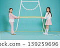 Two girls stand on a blue floor holding a long rope between them. They look happy as they prepare to play a game. The background is bright and simple which focuses attention on them. 139259696