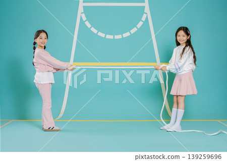 Two girls stand on a blue floor holding a long rope between them. They look happy as they prepare to play a game. The background is bright and simple which focuses attention on them. Two girls stand on a blue floor holding a long rope between them. They look happy as they prepare to play a game. The background is bright and simple which focuses attention on them. 139259696