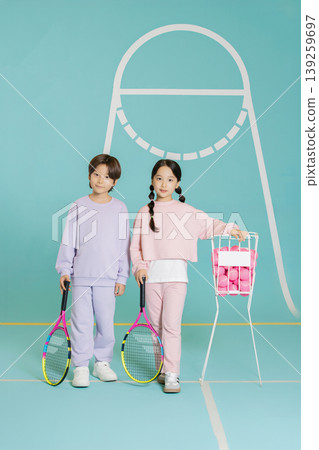 Two kids are on a tennis court. One child holds a racket and a basket of tennis balls. The other child also has a racket in hand. They look ready for practice. Two kids are on a tennis court. One child holds a racket and a basket of tennis balls. The other child also has a racket in hand. They look ready for practice. 139259697