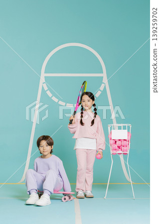 Two children engage in tennis practice at a colorful sports area. One child holds a racket while the other sits on the floor. They enjoy their time with tennis balls. 139259702
