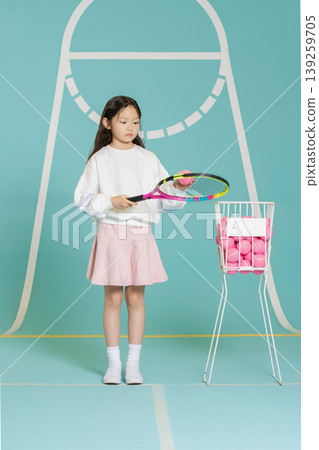 A girl stands on a blue tennis court holding a racket. She looks ready to play. Next to her, there is a basket filled with pink tennis balls. This scene shows her focus on the sport. A girl stands on a blue tennis court holding a racket. She looks ready to play. Next to her, there is a basket filled with pink tennis balls. This scene shows her focus on the sport. 139259705
