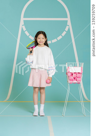 A child is in a tennis court setting. She holds a racket in one hand and a pink ball in the other. A basket with pink balls is next to her. The background is bright and teal. 139259709