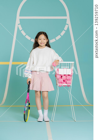 A young girl stands on a tennis court holding a racket in one hand and a ball in the other. A basket filled with tennis balls is beside her. She wears a simple outfit and smiles. A young girl stands on a tennis court holding a racket in one hand and a ball in the other. A basket filled with tennis balls is beside her. She wears a simple outfit and smiles. 139259710