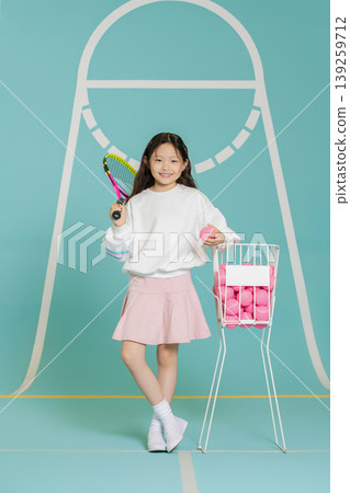 A girl stands with a tennis racket in one hand and a pink ball in the other. She smiles in front of a light blue tennis court. A basket holds more pink tennis balls nearby. A girl stands with a tennis racket in one hand and a pink ball in the other. She smiles in front of a light blue tennis court. A basket holds more pink tennis balls nearby. 139259712