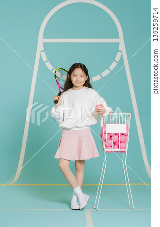 A young girl stands with a tennis racket in one hand and a pink ball in the other. She is dressed in a light outfit and is next to a basket filled with tennis balls. A young girl stands with a tennis racket in one hand and a pink ball in the other. She is dressed in a light outfit and is next to a basket filled with tennis balls. 139259714