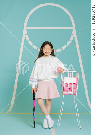 A young girl stands with a tennis racket in one hand and a pink tennis ball in the other. She smiles while next to a cart filled with pink tennis balls. The background is bright and playful. A young girl stands with a tennis racket in one hand and a pink tennis ball in the other. She smiles while next to a cart filled with pink tennis balls. The background is bright and playful. 139259715