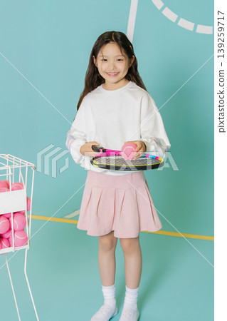A young girl stands in a sports area with a tennis racket in one hand and a pink tennis ball in the other. She smiles and wears a simple outfit with a skirt. A young girl stands in a sports area with a tennis racket in one hand and a pink tennis ball in the other. She smiles and wears a simple outfit with a skirt. 139259717