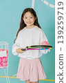 A girl stands in a bright space holding a tennis racket in one hand and a pink tennis ball in the other hand. She wears a white top with a pink skirt and smiles happily. 139259719