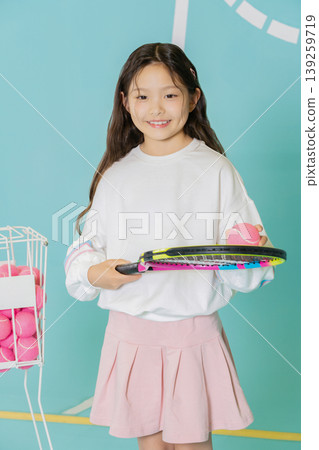 A girl stands in a bright space holding a tennis racket in one hand and a pink tennis ball in the other hand. She wears a white top with a pink skirt and smiles happily. A girl stands in a bright space holding a tennis racket in one hand and a pink tennis ball in the other hand. She wears a white top with a pink skirt and smiles happily. 139259719