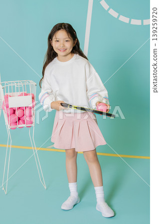 A girl practices tennis on a colorful court. She holds a racket in one hand and a bright pink ball in the other. A basket of pink tennis balls is seen beside her. She is focused and ready. A girl practices tennis on a colorful court. She holds a racket in one hand and a bright pink ball in the other. A basket of pink tennis balls is seen beside her. She is focused and ready. 139259720