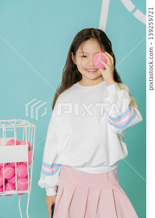 A child holds a pink tennis ball to her eye and smiles. She stands beside a wire basket filled with more pink balls. The background shows bright colors, creating a fun atmosphere. 139259721