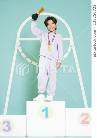 A child stands on the first place podium wearing a medal and holding a trophy high. The background features colorful numbers indicating places in a sports event. The setting looks playful and bright. 139259722