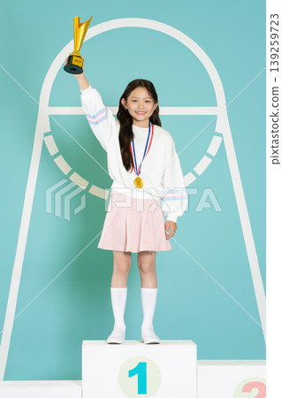 A young girl stands on a podium holding a trophy high with a big smile. She wears a medal around her neck and a sports outfit. The background shows a simple design for an event. A young girl stands on a podium holding a trophy high with a big smile. She wears a medal around her neck and a sports outfit. The background shows a simple design for an event. 139259723