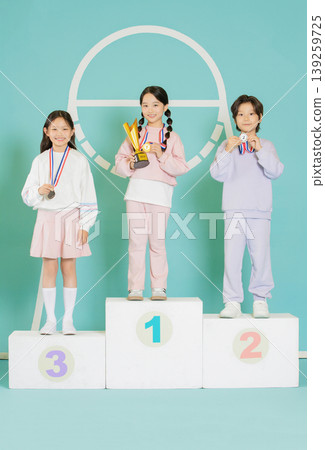 Three children stand on a podium, smiling and holding medals and trophies. They display their awards in a competition setting. The background is bright and colorful, adding to the joyful atmosphere. Three children stand on a podium, smiling and holding medals and trophies. They display their awards in a competition setting. The background is bright and colorful, adding to the joyful atmosphere. 139259725