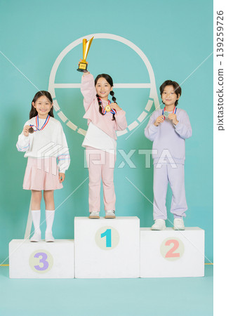 Three kids stand on a podium celebrating their success. One child holds a trophy high, while others show their medals. The backdrop is colorful and cheerful. Three kids stand on a podium celebrating their success. One child holds a trophy high, while others show their medals. The backdrop is colorful and cheerful. 139259726