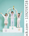 Three children stand on a podium, smiling and raising their hands. Each child wears a medal. One child holds a trophy. The scene takes place in a light-colored room. 139259728