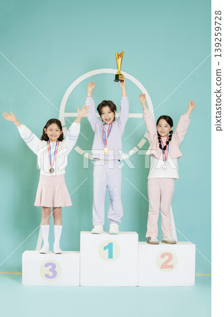 Three children stand on a podium, smiling and raising their hands. Each child wears a medal. One child holds a trophy. The scene takes place in a light-colored room. Three children stand on a podium, smiling and raising their hands. Each child wears a medal. One child holds a trophy. The scene takes place in a light-colored room. 139259728