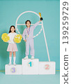 Two children stand on a podium after winning a competition. One child holds a trophy, while the other displays pom poms. The setting is bright and colorful, highlighting their achievements. 139259729